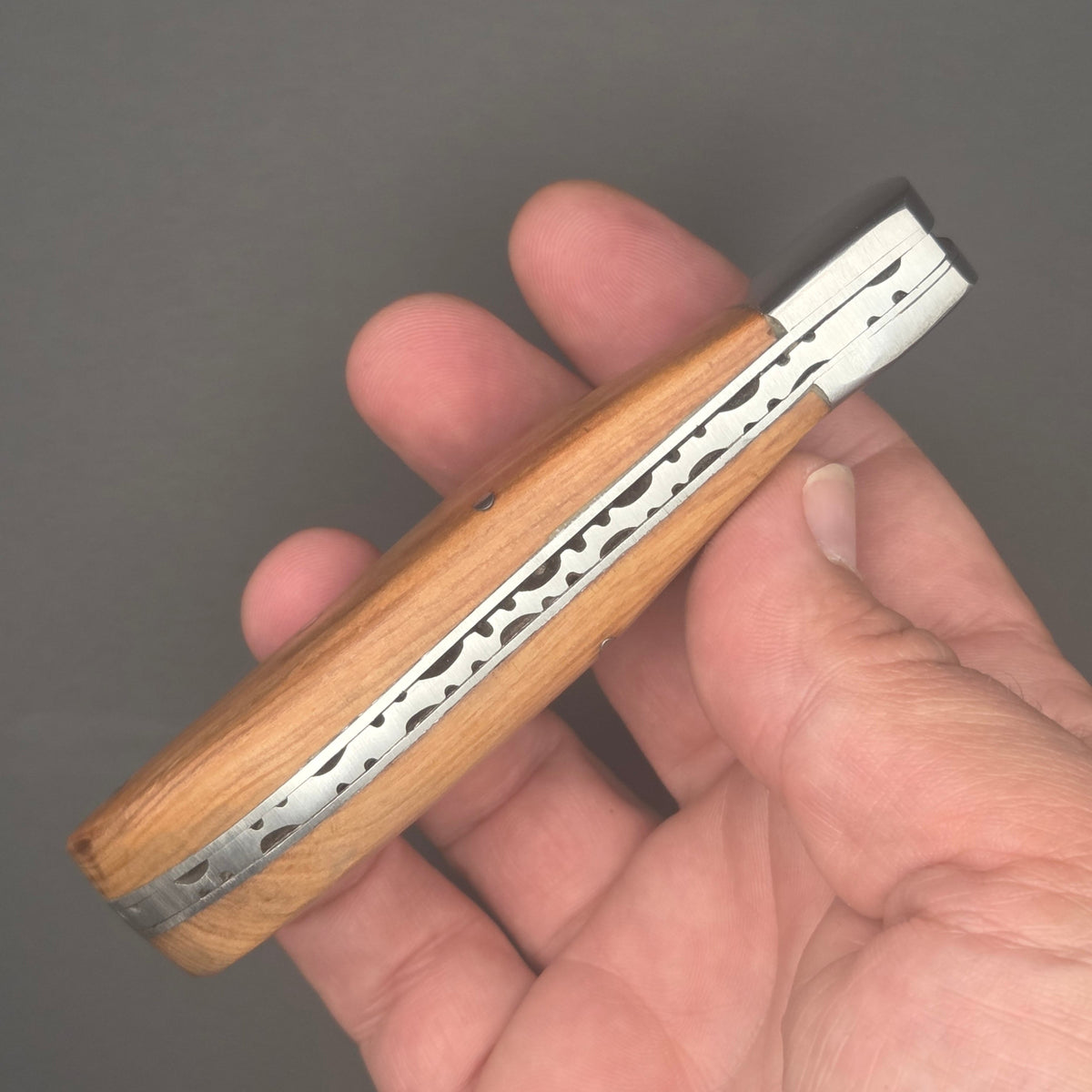 vue du guillochage d'un couteau utilitaire pliant robuste muni d'un manche en teck. Le couteau est tenu dans une main en position fermée. La qualité est de second choix.