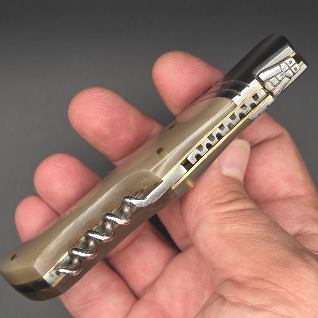 couteau pliant utilitaire tenu en main en position pliée pour contempler le guillochage et le tire-bouchon. ce couteau est muni d'une lame damas