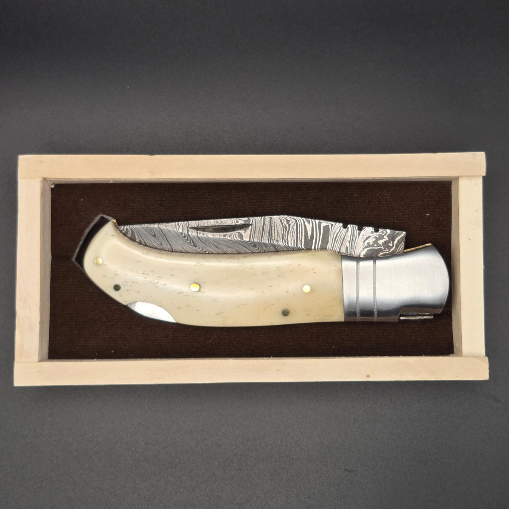 couteau utilitaire pliant en position fermée installé dans sa belle boîte en bois. Le couteau a une lame en damas et un manche en os