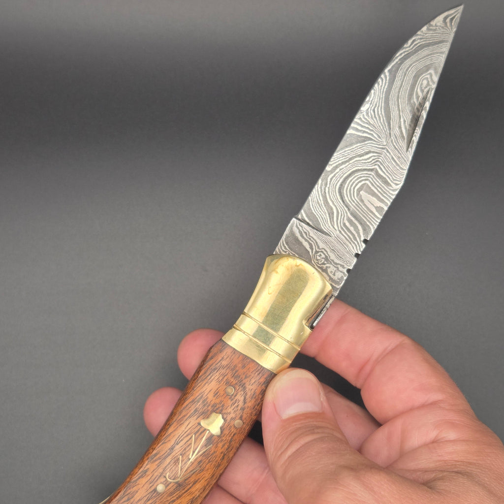 couteau utilitaire pliant avec une lame en damas en position ouvert tenu dans une main. Le manche est en noyer avec une fleur incrustée