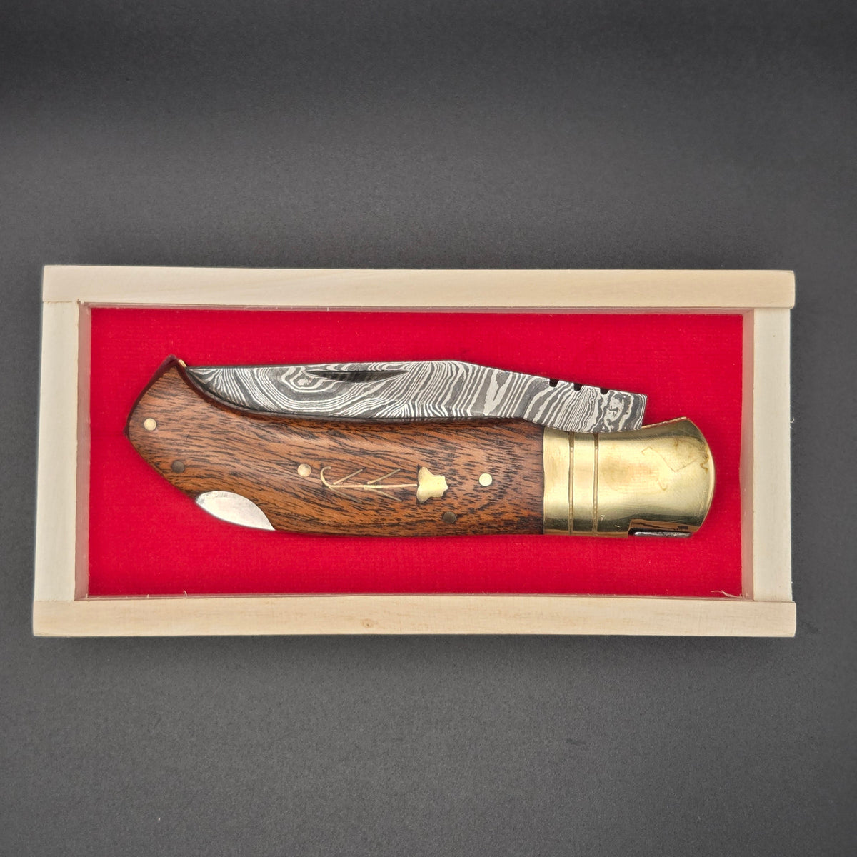 couteau utilitaire pliant avec un manche en noyer dans lequel est incrusté une illustration de fleur possédant une lame en damas présenté dans son coffret en bois