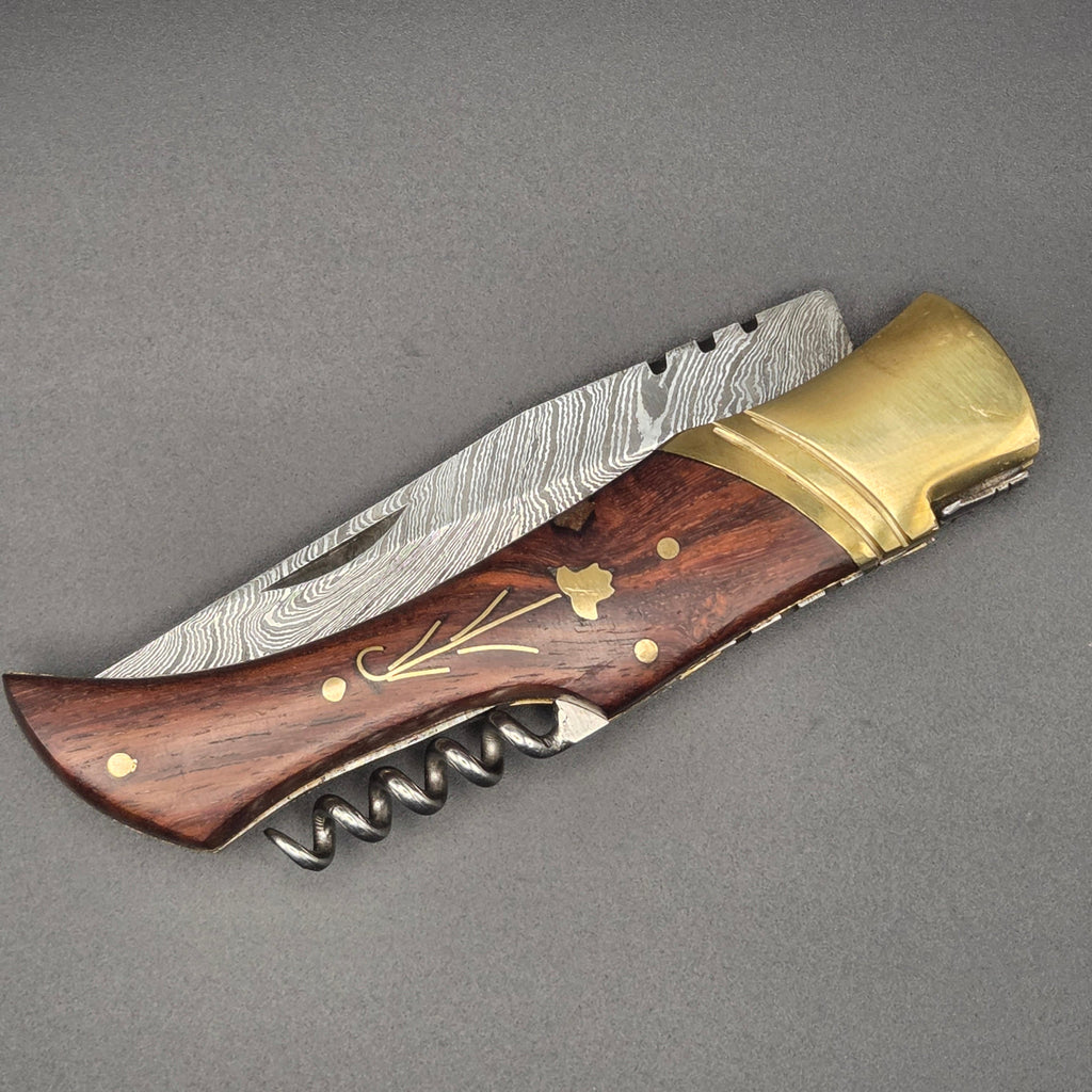 couteau pliant utilitaire avec lame damas et manche en noyer incrusté d'une fleur. le couteau est en position fermée et on y voit le tire-bouchon comme accessoire