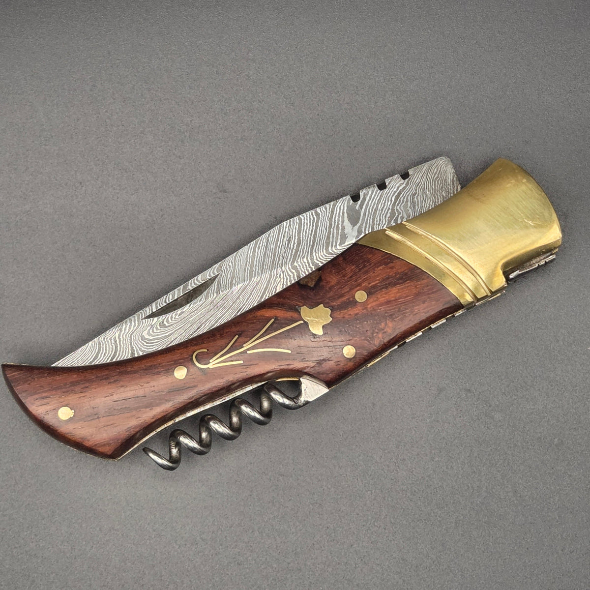 couteau pliant utilitaire avec lame damas et manche en noyer incrusté d'une fleur. le couteau est en position fermée et on y voit le tire-bouchon comme accessoire
