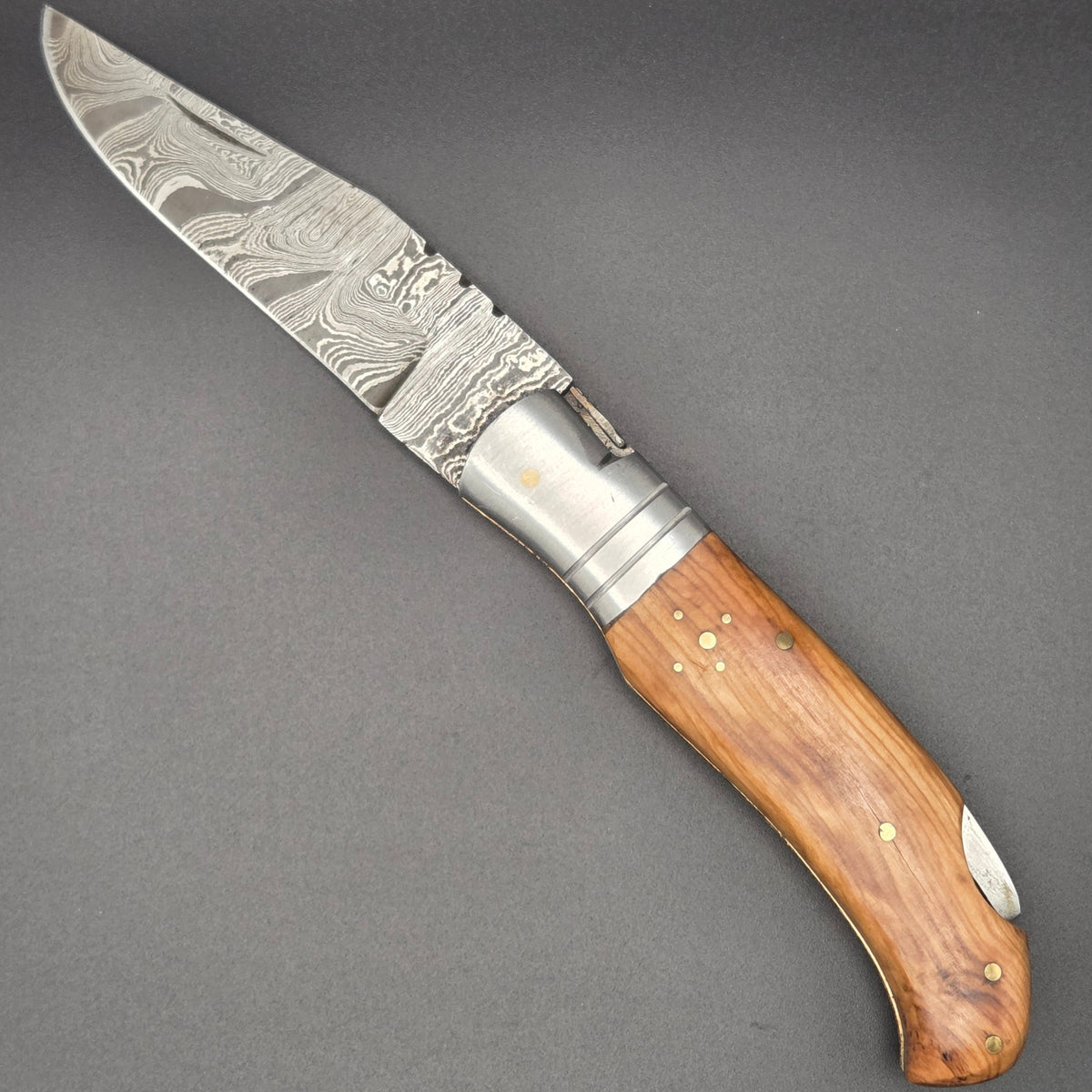 Couteau utilitaire pliant avec lame damas en position ouvert posé à plat. Il possède un manche en teck.