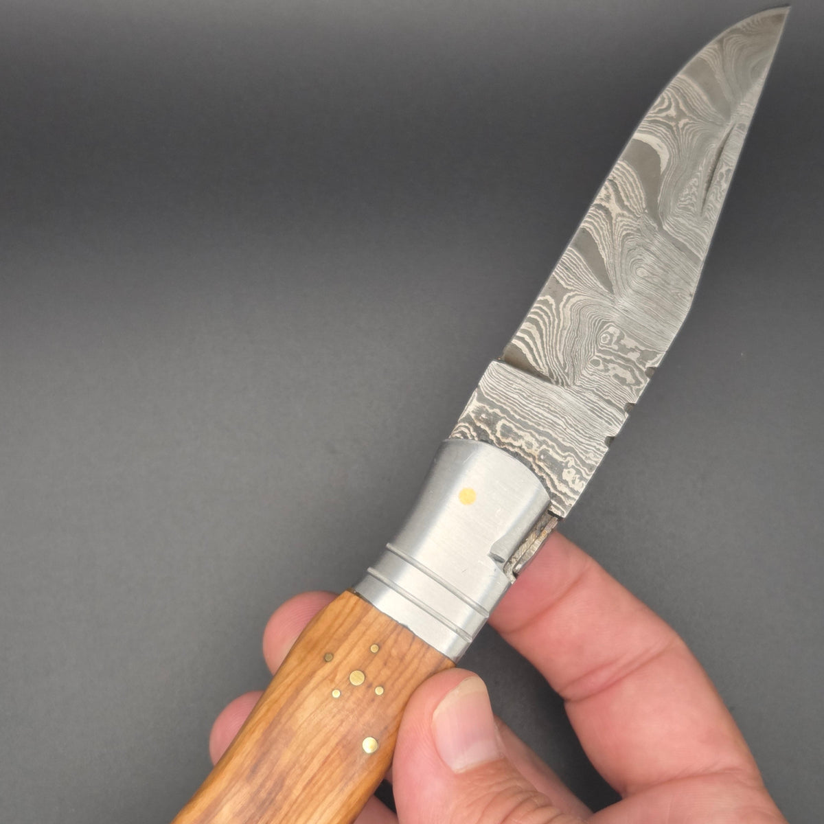 couteau pliant utilitaire en position ouvert et tenu dans une main. Il a une lame en damas et un manche en teck.
