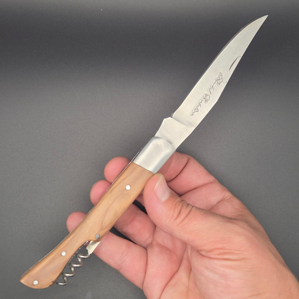 Couteau utilitaire pliant tenu dans une main en position ouvert. Il a un manche en teck et un tire-bouchon