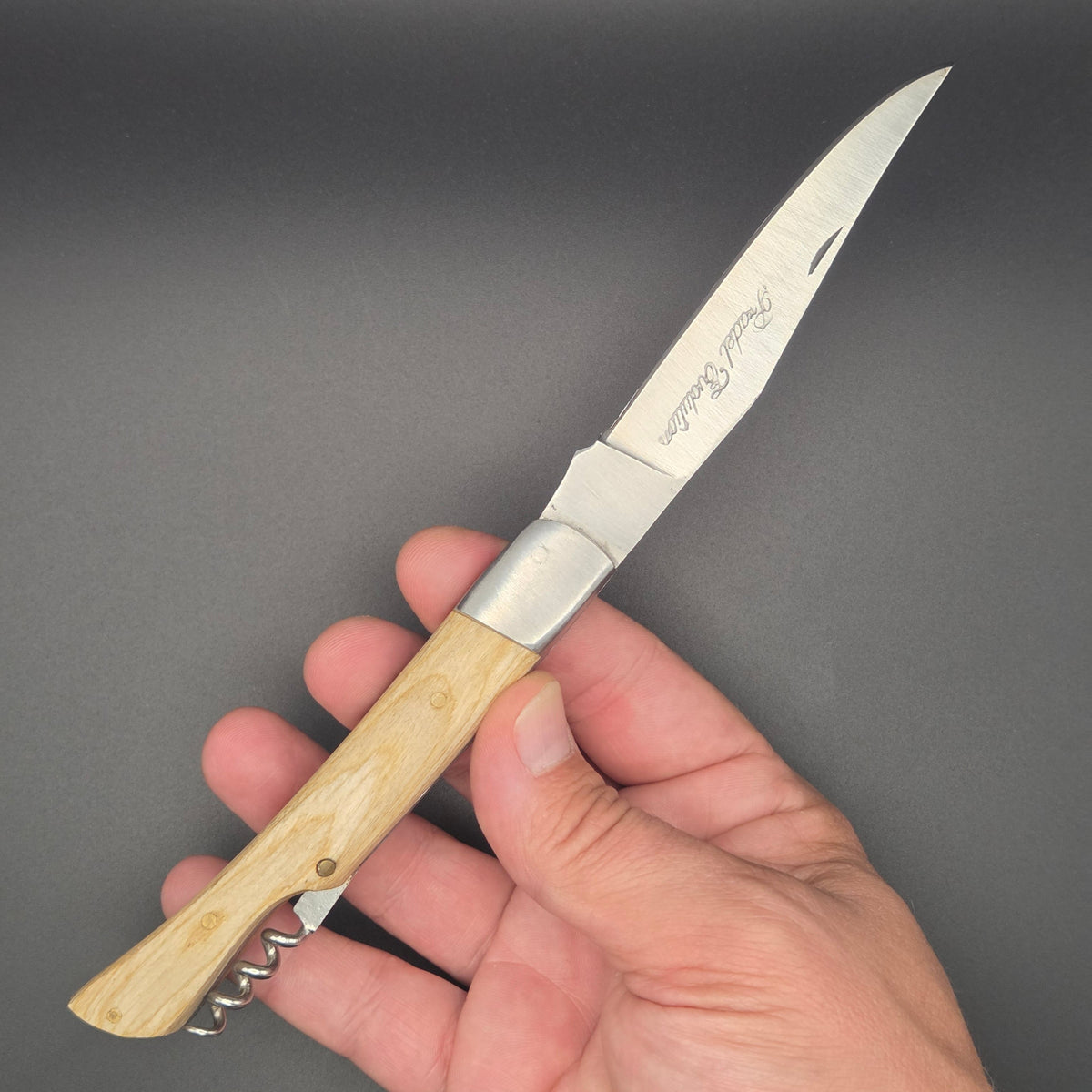 Couteau utilitaire pliant avec un manche en teck blanchi tenu dans une main en position ouvert. Il est muni d'un tire-bouchon.