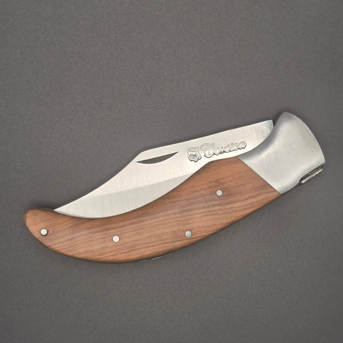 Couteau utilitaire pliant muni d'un manche incurvé en teck. On voit l'indication Corsica gravée sur la lame. Il est posé à plat en position fermé.
